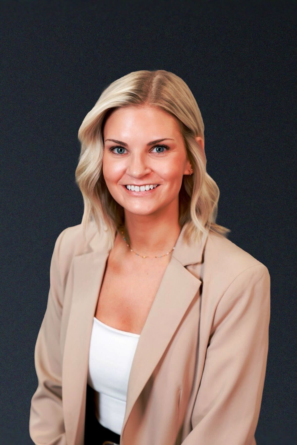 A woman with blonde hair wearing a beige blazer and white top, smiling in front of a plain dark background.