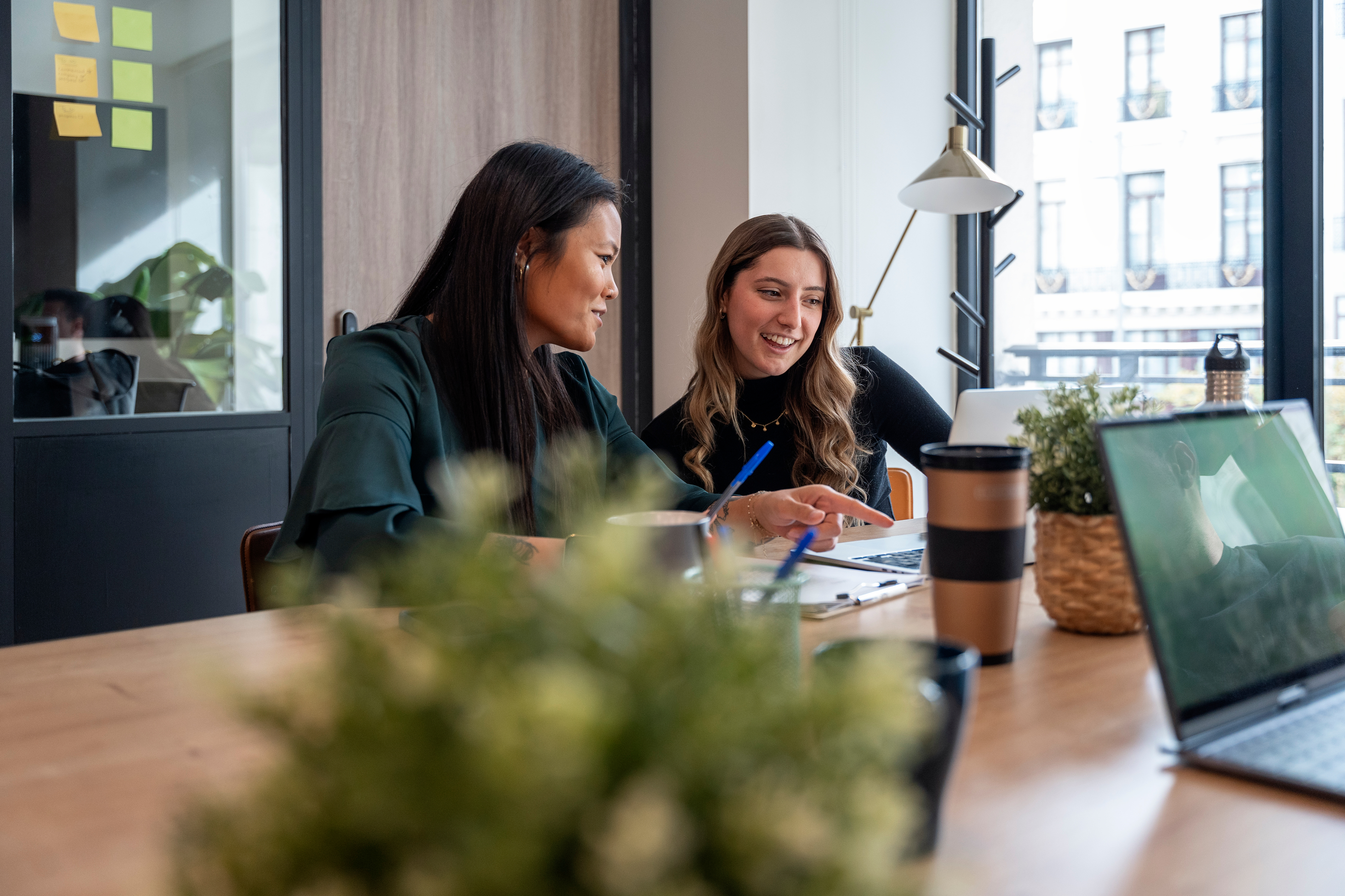 Two women sit at a desk, looking at documents and a laptop, engaged in discussion. Office plants, a coffee cup, and notebooks are on the table.
