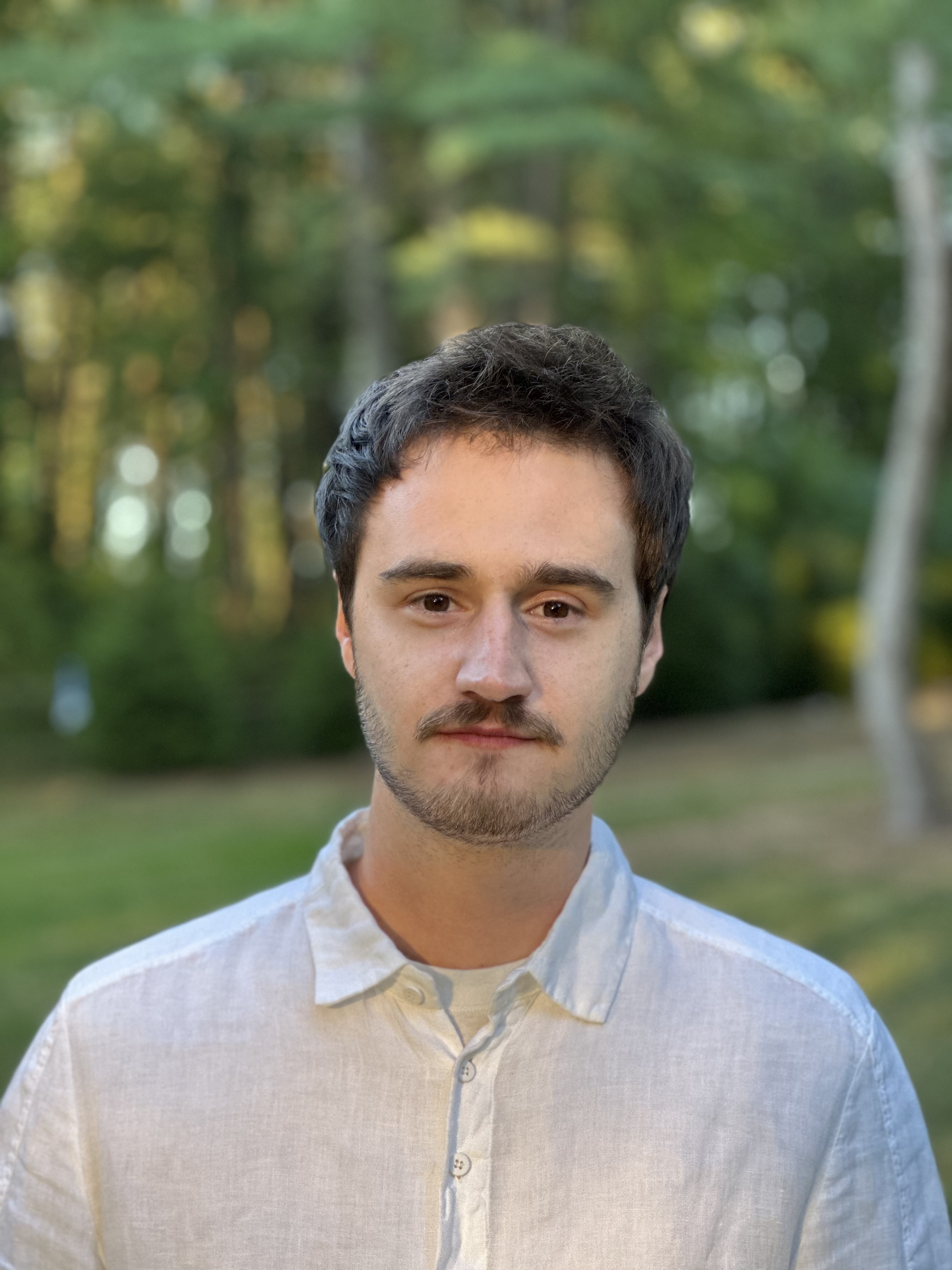 Young man with short brown hair and a mustache, wearing a light-colored button-up shirt, stands outdoors with trees and greenery in the background.