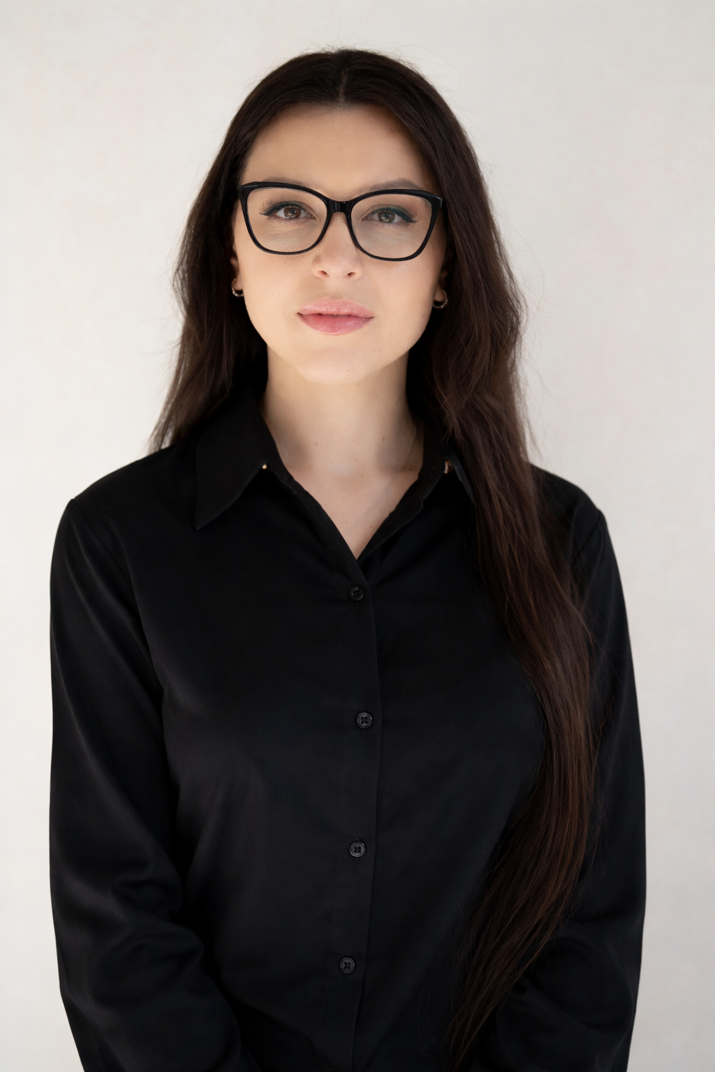 Woman with long dark hair and glasses, wearing a black button-up shirt, stands against a plain light background, looking directly at the camera with a neutral expression.