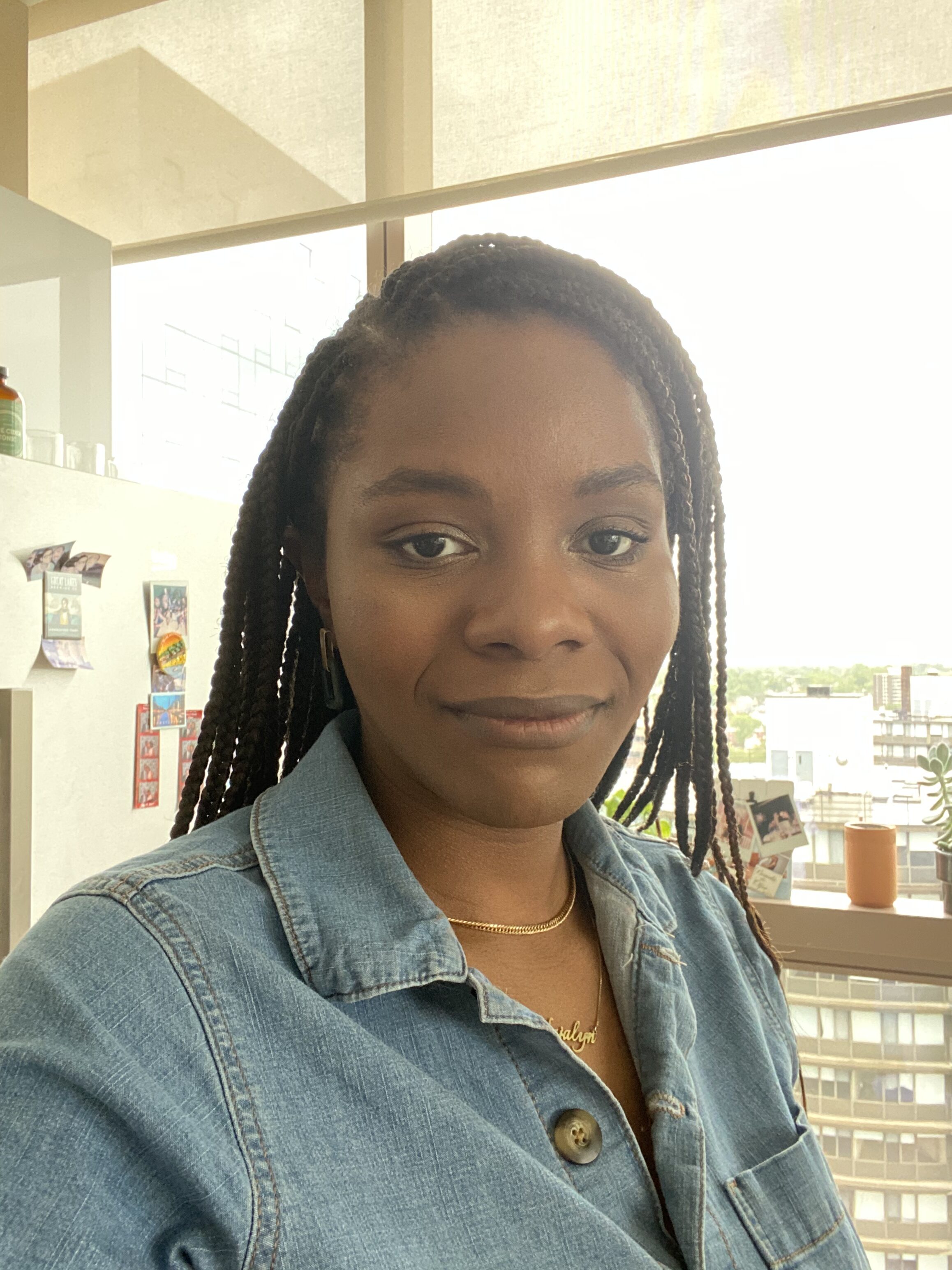 A woman with long braids wearing a denim shirt and gold necklace stands indoors in front of a window and a shelf with photos and plants.