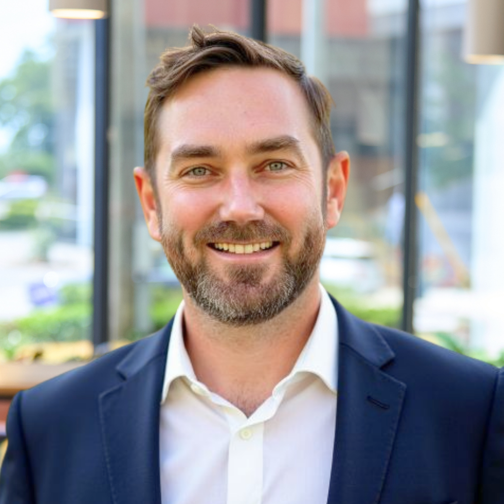 A man with short brown hair and a beard, wearing a white shirt and dark suit jacket, smiles at the camera in an outdoor setting.