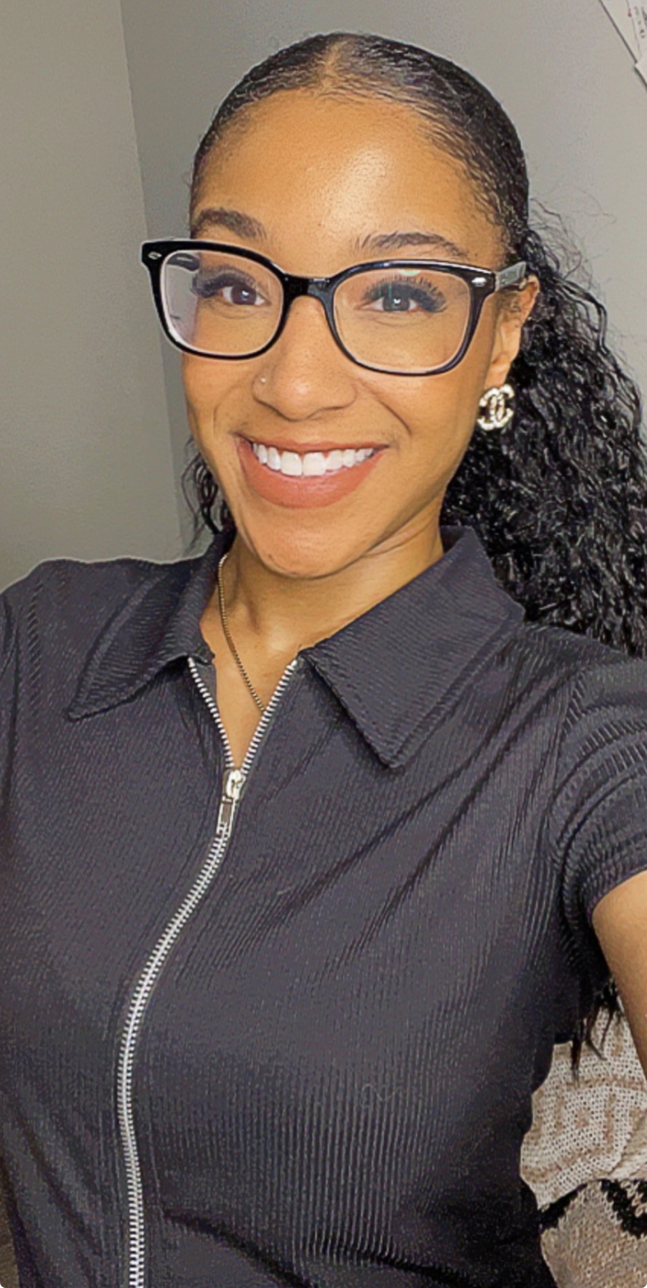 Woman wearing glasses and a black zip-up collared top, smiling at the camera in an indoor setting.