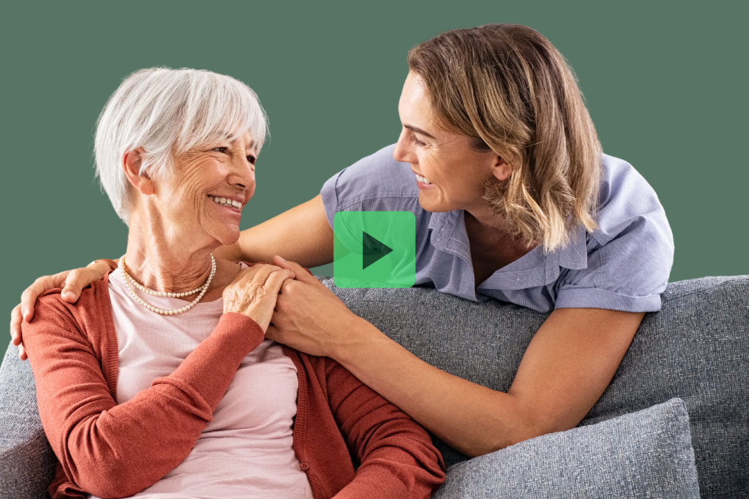 Two women sitting on a couch, smiling at each other. One has gray hair and wears a cardigan, the other has light brown hair and a collared shirt. A play button is overlaid on the image.