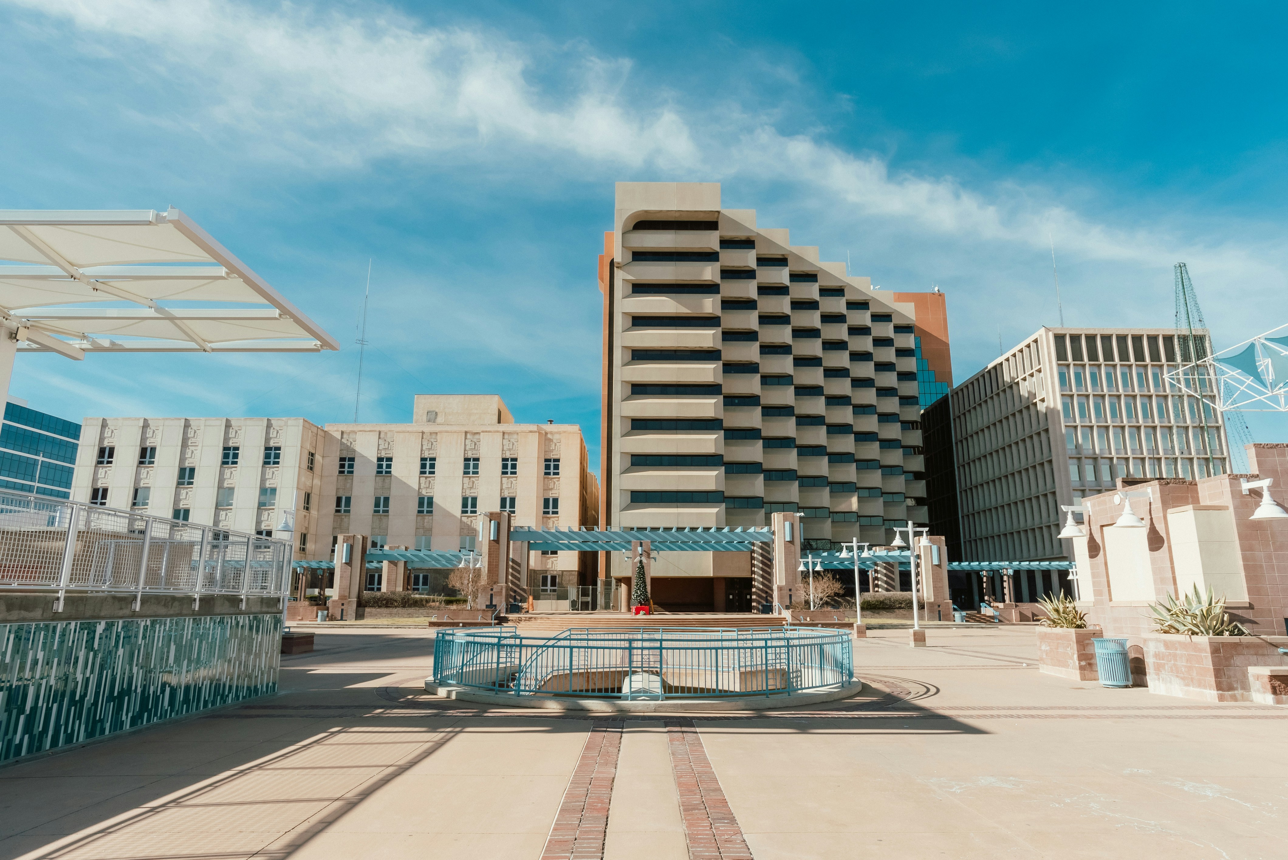 Modern urban plaza with geometric concrete buildings and a blue sky in the background, featuring architectural details and outdoor seating areas.