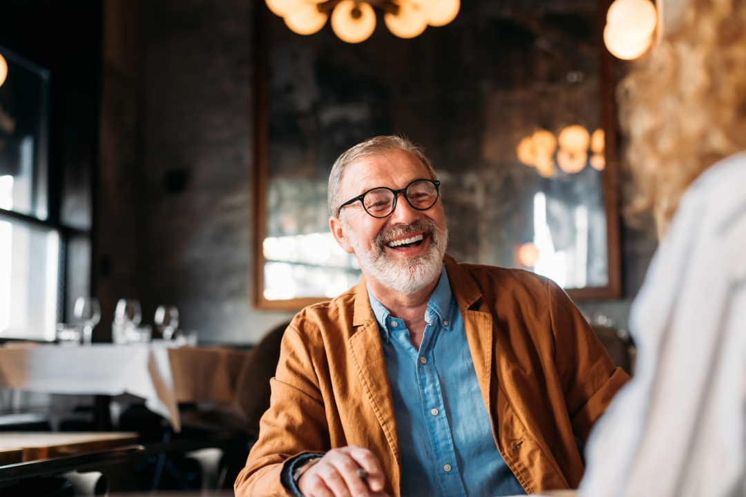 Older man with gray hair and beard, wearing glasses, a blue shirt, and brown jacket, smiling while sitting at a table in a warmly lit restaurant.