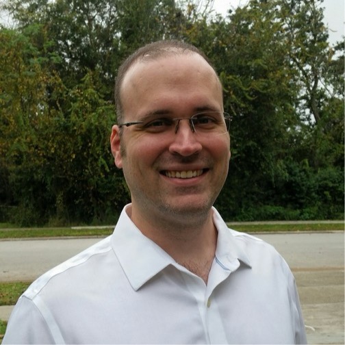 A man wearing glasses and a white collared shirt stands outdoors, smiling, with trees and a road in the background.